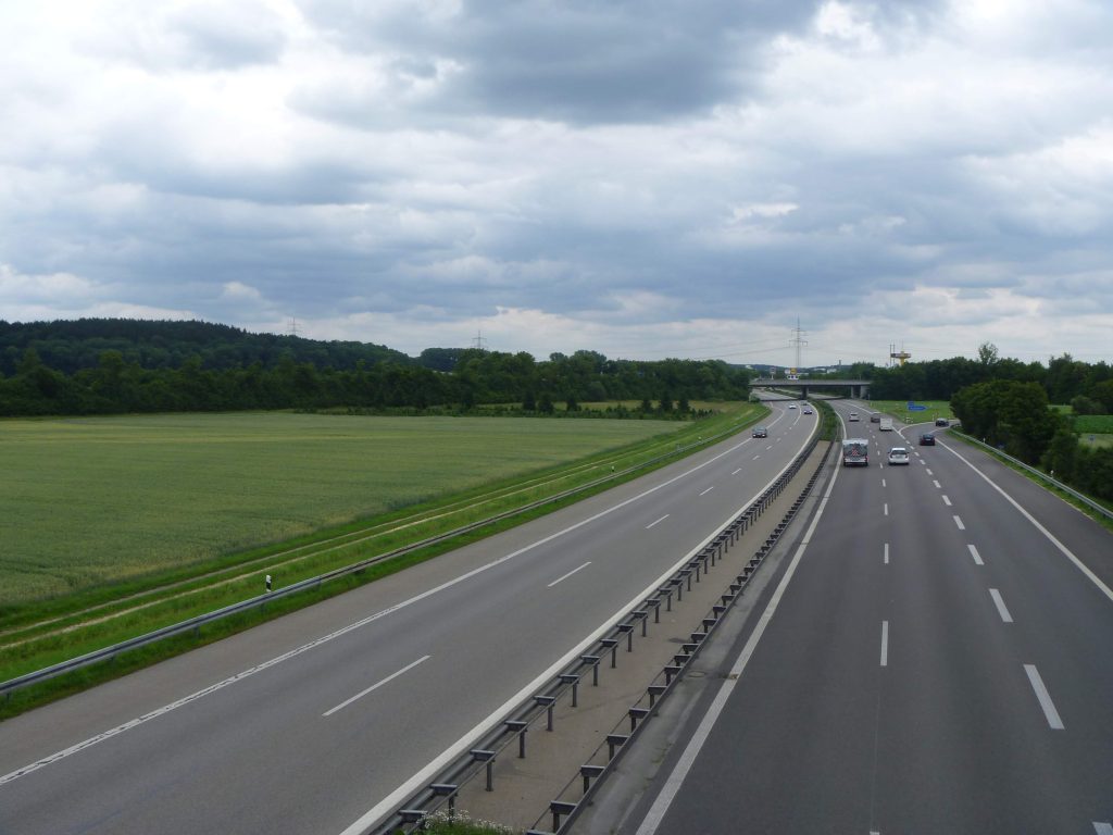 Service-1-min (1) A serene highway scene with cars, green fields, and an overcast sky.